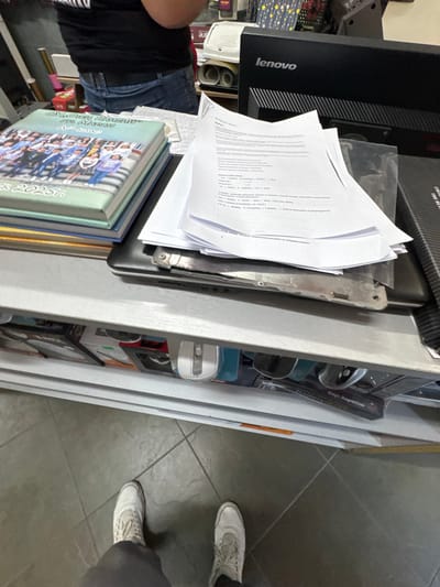The image shows a close-up, overhead view of a desk cluttered with various items. In the foreground, a stack of books with colorful covers is visible, one of which has the text "MR. MAPAN 2025" on its spine. To the left, a black Lenovo monitor is present. Several white sheets of paper are spread across the desk, some appearing to be documents or notes. Also on the desk are what look like parts of a disassembled laptop, a computer mouse, and a box with text that includes "Magic Magnetism" and "Battery 255". The overall scene suggests a workspace or a desk that is in use, possibly for a repair or project. The lighting is bright, indicating daytime, and the tiled floor beneath the desk is visible. The presence of shoes in the lower right corner suggests someone is standing at the desk. The location is identified as Madan, Bulgaria.