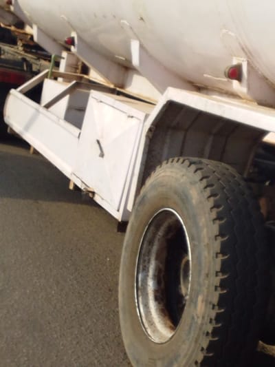 A partial view of a tanker truck is captured from a low angle, showing the white, rounded tank and a side compartment. A large, dirty tire with a silver rim dominates the foreground. The tire has deep treads and appears to be well-worn. The vehicle is parked on dark asphalt. The lighting is bright, suggesting daytime.