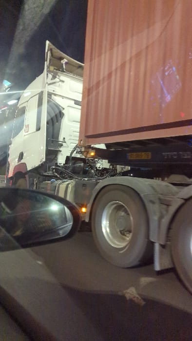This nighttime image captures a white semi-trailer truck and its attached reddish-brown shipping container from the perspective of an adjacent vehicle, on a road in the city of Batzra, Israel.

The truck's cab, positioned on the left side of the frame, is white with an upward-angled roof spoiler/air deflector, appearing partially open at its top edge. Below the cab, mechanical components including air lines, hoses, and what appear to be fuel tanks are visible. Several amber marker lights on the truck's chassis are illuminated. The truck's rear wheels, with silver hubs, are visible, suggesting a multi-axle configuration. The attached trailer carries a large, reddish-brown shipping container, its side reflecting ambient light sources in abstract patterns. A yellow Israeli license plate, partially legible as "19-978" (or similar digits), is affixed to the trailer's frame.

In the lower-left foreground, a dark side mirror from the camera's vehicle is visible, reflecting distorted light and a faint outline of the interior. The road surface is dark asphalt, with a small, light-colored piece of debris lying near the truck's wheels. The background is dark, with blurred artificial lights indicating other vehicles or infrastructure. No people are visibly present in the scene. The truck appears to be either stationary or in slow parallel motion relative to the camera's vehicle.