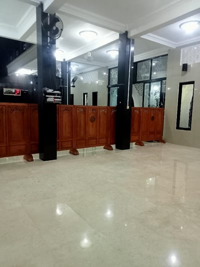 The image shows the interior of a mosque in Kecamatan Margorejo, Indonesia, with a white marble floor. The room is empty with a wooden partition in the foreground, and two black pillars in the background. The mosque is well-lit with multiple ceiling lights and windows. There is a large window with decorative glass and a smaller window with a patterned white film. A ceiling fan can be seen in the top left corner. The room is clean and empty, with no people present.