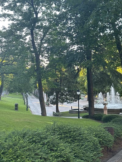 The image shows a pathway in a park, likely in Oviedo, Spain, with lush green grass and trees. The pathway is paved with grey and white tiles and is lined with lampposts. To the right of the pathway, a fountain is spraying water. Several green benches are visible along the pathway and near the fountain. A dark-colored trash can is located on the grass to the left of the pathway. The scene appears to be a public park or garden, with a focus on landscaping and recreational amenities. The time of day is likely late afternoon or early evening, as indicated by the soft lighting.