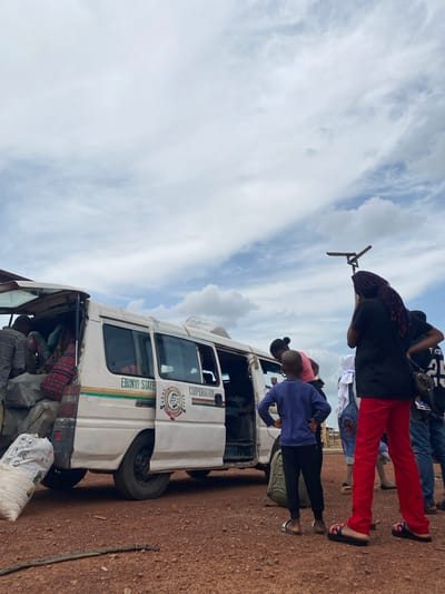 The scene depicts a public transportation van in Abakaliki, Ebonyi State, Nigeria. A group of people are gathered near the van, with some individuals entering and exiting the vehicle. Passengers and belongings are near the van. The van is painted white with green stripes and features the inscription "Ebonyi State Corporation." The weather appears sunny. The ground around the van is dirt.