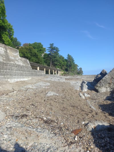 The image depicts a rocky shore with a clear blue sky and some scattered white clouds. The foreground is dominated by a grey, weathered concrete structure with a textured surface of small stones. To the left of the concrete structure, there is a lush green hillside covered with trees. A partially visible set of wooden stairs or a ramp leads up the hillside. The overall scene suggests an outdoor setting, possibly a coastline or a body of water. The time of day appears to be daytime, given the bright sunlight and clear sky. There is no visible text in the image. The orientation of the image is tilted.