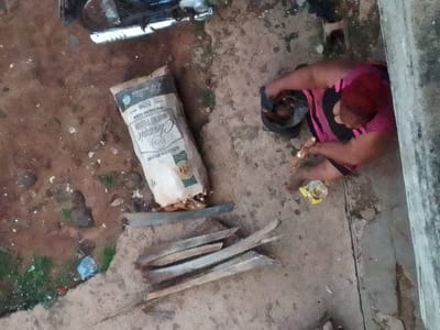The image appears to be taken in a public or semi-public outdoor space, possibly in Orlu, Nigeria. The scene includes a woman in a pink and black striped top kneeling next to a concrete surface. She is near a black bag, possibly containing food items. Next to her is a yellow package of snacks and a bag of "Classic Wheat Flour for Baking 50KG, Golden Penny" which is partially torn and spilling contents. There are shards of wood or sticks are scattered near the flour bag. In the upper portion of the image, there is a partial view of a motorcycle. The ground is a mix of dirt, concrete, and some vegetation. The light is somewhat dim and overcast.