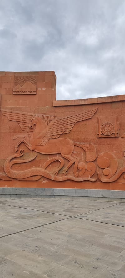 The image captures a large, brick-red relief carving on a wall. The main subject is a winged horse, similar to Pegasus, in a dynamic pose, seemingly attacking a serpent or dragon-like creature coiled beneath it. To the upper left of the horse is a smaller, rectangular carving featuring a mountain landscape, possibly Mount Ararat, with Armenian script underneath. To the upper right of the horse is a circular emblem or crest, flanked by vertical rectangular bars, possibly swords or markers. The wall is made of brick-red rectangular blocks, and there is a stone-paved area in the foreground, suggesting a public space. The sky above is overcast. This image shows Araks in Armenia.