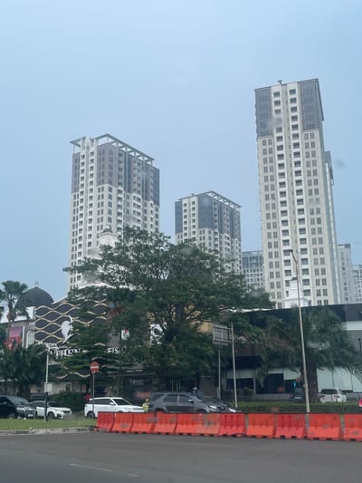 The image captures a street view in Pakulonan Barat, Indonesia, dominated by modern high-rise apartment buildings under an overcast sky. In the foreground, orange construction barriers line a road, with several cars parked and driving. A person wearing a yellow vest stands near a white SUV. Behind the road and barriers, a multi-story building with a black wavy facade and a dome structure is visible, partially obscured by trees. Further back, several tall apartment buildings with a contemporary design, featuring numerous windows and grid-like facades, rise into the sky. The overall scene suggests an urban environment, potentially during or after a period of rain, given the muted colors and slight haze.