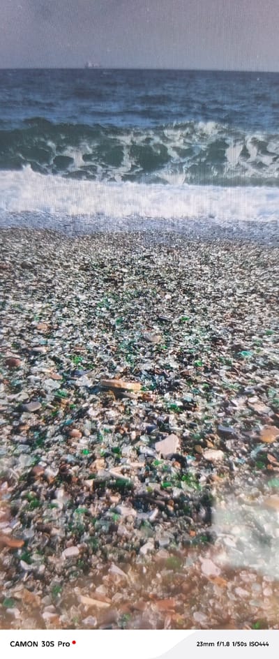 The image displays a close-up view of a beach covered in a multitude of glass fragments of various colors, including green, brown, and clear. In the background, the sea is visible with gentle waves breaking on the shore, and a hazy, distant landmass or ship can be seen on the horizon under a gray sky. The foreground is densely packed with the polished glass pieces, suggesting a unique beach environment. The text overlay indicates "CAMON 30S Pro" and "23mm f/1.8 1/50s ISO444", which are likely camera settings and model information. This scene is representative of Glass Beach in Nakhodka, Russia, a location known for its transformation from a dumping ground to a unique beach of sea-tumbled glass. There are no people or animals present in the image.