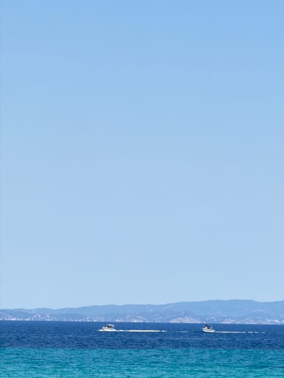 This is a vertical, eye-level, medium shot of a bright, clear blue sky dominating the left side of the frame. On the right side, the tranquil, deep blue sea meets a distant, hazy mountain range. A white sailboat is visible on the water, moving away from the viewer. The sun is high, indicating it's midday. The weather appears to be clear and fair, perfect for sailing. The scene evokes a sense of peace and freedom, with the vastness of the sky and sea offering a sense of escape. The location is Sithonia, Greece, known for its beautiful coastlines and mountainous terrain. There is no visible text in the image.