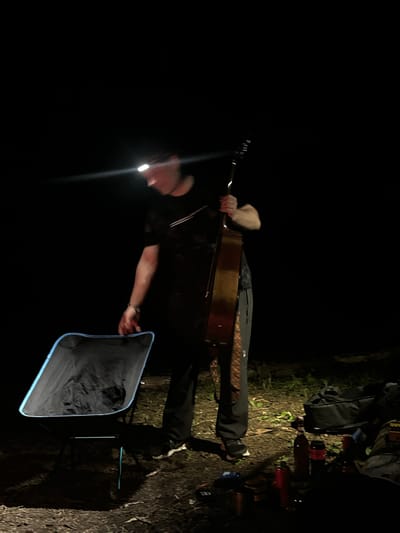 A person wearing a headlamp stands in a dark, outdoor environment and holds a guitar. A folding chair is beside them, and a backpack and various items are on the ground nearby. The location appears to be at night in a rural or wilderness setting, consistent with Юзуфоўскі сельскі Савет, Belarus.
