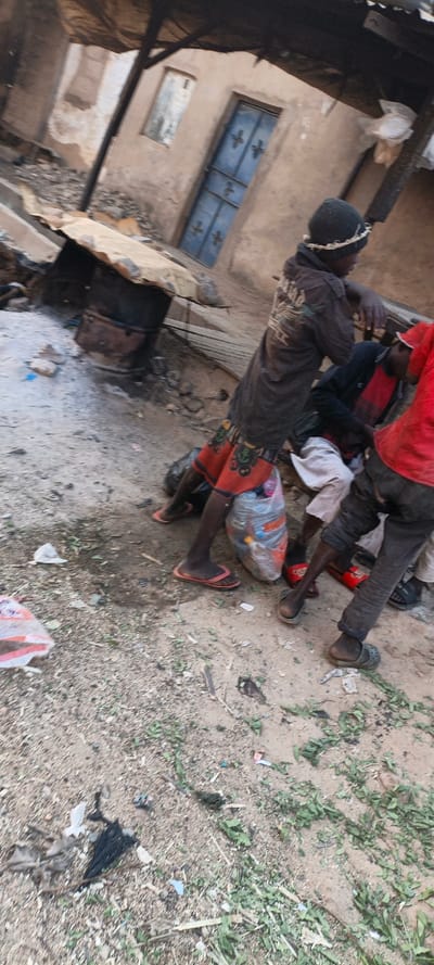 Amidst a dilapidated landscape with damaged buildings, a group of children gathers. One wears a beanie, a dark jacket, and red patterned shorts, holding a plastic bag filled with various items. Another child sports a red shirt and dark pants, while a third is partly visible. The ground is littered with debris and sparse vegetation, adding to the overall impression of poverty and neglect.