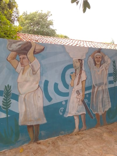 The image depicts a mural located in the city of Villa Hayes, Paraguay. The mural features three painted figures of people carrying items on their heads. All three figures are wearing light-colored, knee-length garments.

The figure on the left is carrying a gray, stone-like object. The figure in the middle has dark hair braided and is carrying a long, dark object on their left side. The figure on the right has their hands raised to their head and appears to be carrying something under a cloth.

The background of the mural is a light blue color with abstract, curved shapes. Green leaf-like shapes are painted beside the figures. The mural is painted on a wall topped with red roof tiles. Parts of green trees and foliage are visible above the roof tiles. The ground in front of the mural is a beige, textured surface.