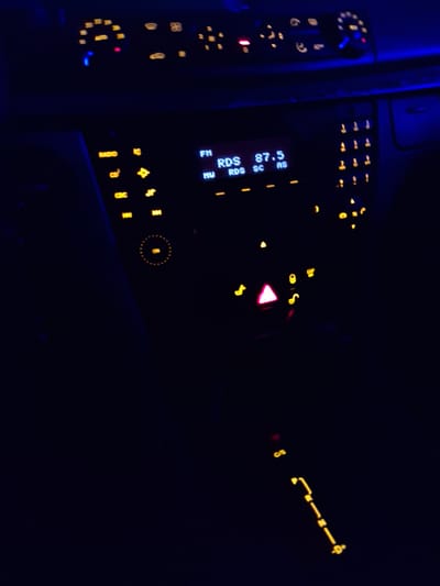 In the dimly lit interior of a car, the dashboard displays illuminated controls and indicators, suggesting it is night or the interior lights are the primary source of illumination. The scene is set in Piilo, Ukraine, though the specific location within the city is not discernible from the image alone. The primary focus is the car's dashboard, featuring a digital display for the radio showing "RDS 87.5" and "RDS SC AS". Various buttons and controls are also lit, including those for radio functions like "RADIO" and "CD", and an "ON" indicator within a circular light. To the right, a numerical keypad is visible. Above the radio, a speedometer and other indicator lights are dimly lit. Below the radio and extending downwards, a gear selector is illuminated, showing the positions "P", "R", "N", and "D". A triangular hazard light indicator is also illuminated in pink. No people are visible in the frame.