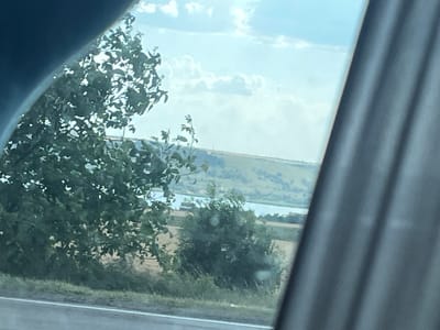 The image depicts a landscape viewed through a car window, suggesting a journey. In the foreground, a large tree with dense green foliage is prominent on the left, partially obscuring the view. To the right, a dark curtain or car interior element frames the scene.

The background reveals a body of water, likely a river or lake, stretching across the middle ground. Beyond the water, rolling hills rise, some appearing cultivated with patches of yellowing fields, suggesting agriculture. The sky above is a soft blue with scattered white, fluffy clouds, indicating fair weather. The lighting suggests it is daytime, possibly late morning or afternoon, with the sun casting a gentle light.

There are no people or explicit objects visible, only natural elements and the implied context of being inside a vehicle. The scene evokes a sense of peaceful travel through a rural or semi-rural Ukrainian landscape. No text is visible in the image.
