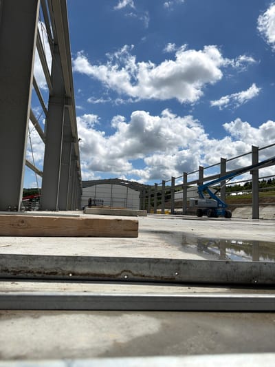 An outdoor, eye-level, medium shot captures a construction site under a bright, cloudy sky in Skrzydlna, Poland. The skeletal structure of a large building, composed of grey steel beams, dominates the left side of the frame. In the mid-ground, a blue aerial work platform, identified by the "Genie" logo, is parked on a wet, concrete surface. A partially constructed building with a white roof is visible in the background, along with rolling, tree-covered hills. In the foreground, a wooden plank rests on the concrete, and the bottom edge of the image shows two parallel metal strips. The scene suggests ongoing construction activity.