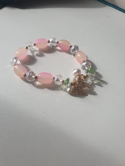 The image displays a single bracelet positioned on a plain, light-colored, flat surface. The bracelet is composed of alternating light pink, opaque, oval-shaped beads and clear, faceted, spherical beads. Attached to the bracelet is a gold-colored, angel-shaped charm embellished with small, reflective elements, alongside one small, white, spherical pearl and several small, green, faceted beads. The bracelet is arranged in an open, roughly circular configuration. Strong, directional illumination from the upper-left creates distinct shadows extending towards the lower-right of the frame. The background is out-of-focus, revealing indistinct dark and faint blue elements in the upper portions. The image contains no discernible visual elements to indicate a specific location within Ulaanbaatar, Mongolia, or any other geographical area.