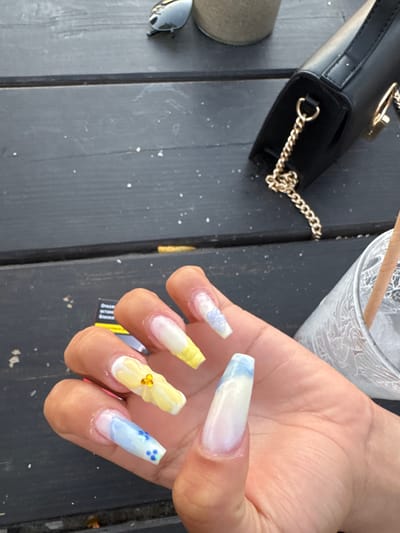 A close-up shot shows a person's hand with long, decorated acrylic nails resting on a dark, weathered wooden surface. The nails feature a gradient of colors, including white, light blue, and yellow, with some adorned with floral designs and small embellishments. To the left of the hand, a black handbag with a gold chain strap is visible. Also on the left, partially obscured, is a pair of dark sunglasses and a neutral-colored object. In the background, a glass with a decorative pattern and a wooden stick are present. The overall setting appears to be outdoors, possibly on a table, with scattered white specks on the wooden surface suggesting debris or an outdoor element. The lighting suggests it is daytime. A small piece of yellow packaging with some text is visible near the fingers. No specific emotions are conveyed.