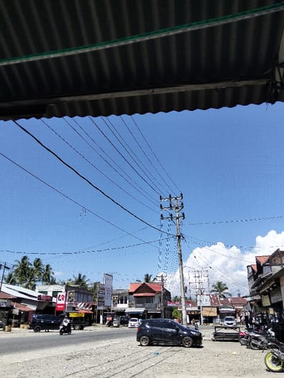An outdoor shot shows a street scene with a focus on utility lines and a pole dominating the clear blue sky. Buildings line the street, featuring a mix of commercial and residential architecture. Vehicles include a dark car, a motorcycle, and parked motorcycles along the roadside. The foreground displays a rough, gravelly texture, contrasting with the smoother road. Overhanging roof structures partially frame the upper portion of the view.