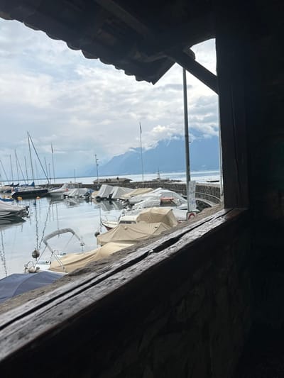 The image depicts a static view of a marina in La Tour-de-Peilz, Switzerland, framed by an interior wooden structure. The foreground features dark, weathered wooden beams across the top and right, with a dark stone wall visible in the lower right, suggesting a window or sheltered opening.
Beyond the frame, the scene focuses on the marina, where numerous boats are moored in calm water. Many vessels are covered with protective tarpaulins in shades of white, beige, and some blue. Several sailboats with visible masts are positioned on the left side of the marina. A stone pier extends into the lake on the right, culminating in a small, white, lighthouse-like beacon structure. A tall, grey utility pole stands vertically near the pier.
In the mid-ground, the expansive waters of Lake Geneva reflect the overcast sky. The background is dominated by a range of large mountains, partially obscured by low-hanging clouds and mist, indicating a cloudy or possibly rainy day. The sky above is entirely overcast with grey clouds. No individuals or active movements are discernible in the scene.