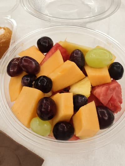 A close-up, overhead shot shows a clear plastic container filled with a colorful fruit salad. The salad contains cubed cantaloupe, chunks of watermelon, and several grapes, some dark purple and some green. The container is resting on a white, textured surface, possibly a tablecloth. In the upper left corner, a portion of another plastic container is visible, containing a golden-brown pastry with a white filling. The lighting is bright and even, highlighting the freshness of the fruit. The background is blurred, drawing focus to the fruit salad. This image is not located in the city of Pozzuoli, Italy.