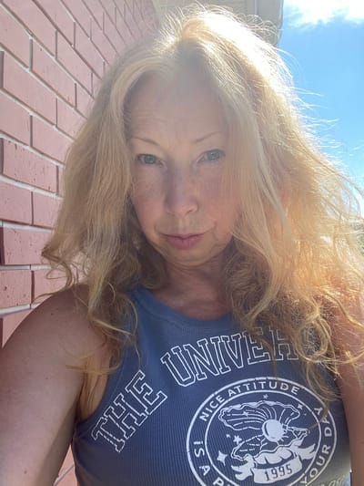 The image is a close-up selfie of a blonde woman with fair skin, taken outdoors against a brick wall and a bright blue sky. She is wearing a grey ribbed tank top with white lettering that says "THE UNIVERSE NICE ATTITUDE IS! A PART OF YOUR DAY 1995". The sunlight is strong, creating highlights in her hair and some glare on her face. Her hair is styled loosely with some curls. The brick wall is pink and appears to be a facade. The overall setting suggests a sunny day in an urban or suburban environment. The location is specified as Bol'shoe Zaborod'e, Russia.