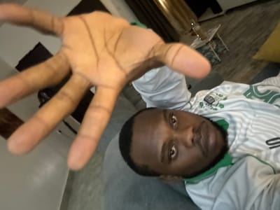 A first-person perspective image shows a man extending his open right hand towards the camera. The man has dark hair and a dark beard. He wears a white shirt with green stripes.

Behind the man and his hand, there is a living room with a grey couch. A small, white, multi-level table is visible, with what appears to be a bottle and a glass on top. A partial view of a yellow pillow is seen.

The location cannot be determined as Iddo Tudunwada, Nigeria based on the image.