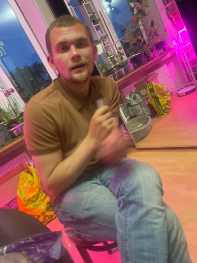 The image shows a young man with short, light brown hair and a light beard, wearing a brown polo shirt and light blue jeans. He is seated on a stool, holding a silver object in his right hand near his mouth. His left arm is bent, resting on his leg. The background features a window with blue sky visible, a shelf unit with plants and pink LED grow lights, and various items on a wooden surface, including yellow bags, a grey pet bed with a yellow toy, and a circular black device. The overall environment appears to be indoors, possibly a living space or a room dedicated to plant cultivation. There are no specific visual cues within the image that would allow for the determination of its location within the city of Vyborg, Russia.