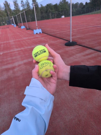In Vantaa, Finland, two individuals' hands are visible on a red tennis court, holding tennis balls. The person in the foreground wears a light blue jacket with the word "rukka" printed in black on the sleeve. Their hand is holding a tennis ball with the branding "SOFTEC SPEED" visible. The other person, whose hand is partially visible in the upper right, is wearing a black jacket and holding another tennis ball. The court is bordered by a net and trees can be seen in the background under a twilight sky.