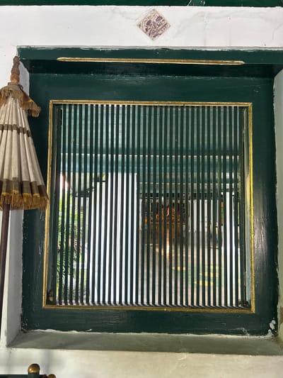 The image shows a close-up view of a green-painted window frame with a gold trim, set against a white wall. The window is covered by a vertical metal grate, also painted green. Through the grate, an interior space is visible, featuring a tiled floor with a yellow and green pattern. There are also glimpses of what appears to be decorative elements, possibly furniture or cultural artifacts, with some red and gold colors. On the left side of the window, a portion of an ornate umbrella or parasol, white with gold tassels and trim, is visible, leaning against the wall. The overall impression is one of a traditional architectural element, possibly within a historical building or palace in Cirebon, Indonesia, a city known for its rich cultural heritage. The lighting suggests it is daytime.
