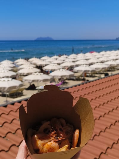 A close-up shot shows a person holding a brown cardboard container filled with fried shrimp and what appears to be potatoes. In the background, a beach is visible with numerous white umbrellas, lounge chairs, and people enjoying the sun. The sea stretches out to the horizon, with a distant island or mountain range faintly visible. The image is taken from a vantage point on a terracotta tiled roof, typical of coastal Italian architecture. The overall impression is of a sunny day at a seaside resort, likely in Sperlonga, Italy, given the architectural style and beach scene.