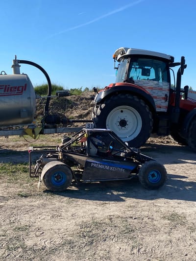 The image depicts a rural scene, possibly a farm or agricultural area. In the foreground, there is a piece of farming equipment labeled "PREMIUM KERÍTÉS" and "31" with "Fliegl RARTECHNIK" visible on a tank. Behind the equipment, a large tractor is partially visible, with markings indicating "STEYR 20 EXPE" and text possibly related to "IPARD" and "max 65%".

The ground is dirt or gravel, and some vegetation is visible in the background. The sky is clear blue with some contrails. The image appears to be taken in Kanjiza Municipality, Serbia, as the label "PREMIUM KERÍTÉS" suggests a Hungarian connection, given the proximity of Kanjiza to Hungary.