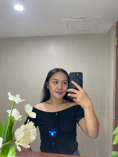 A young woman with long, dark hair is taking a mirror selfie in a bathroom. She is wearing an off-the-shoulder black top and a necklace with a small pendant. She is holding a black smartphone in her right hand, which is angled to capture her reflection. In the foreground, to the left, is a bouquet of white flowers. The bathroom walls are tiled in a light beige color, and the ceiling has a recessed light fixture and an access panel. The overall lighting suggests the photo was taken indoors. This scene is consistent with a personal moment captured within a residential or hotel bathroom in Koronadal City, Philippines.
