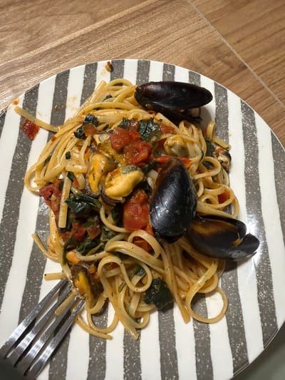 A plate of linguine with mussels and chopped tomatoes sits on a wooden table. A fork is placed to the left of the plate. The plate has a pattern of grey and white stripes. The mussels are dark blue to black with orange-yellow interiors. The linguine is a golden yellow color, coated in a red sauce with visible pieces of tomato and dark green leafy vegetables. The lighting suggests an indoor setting, possibly a home or restaurant. This image does not provide information to determine its location in Catania, Italy.