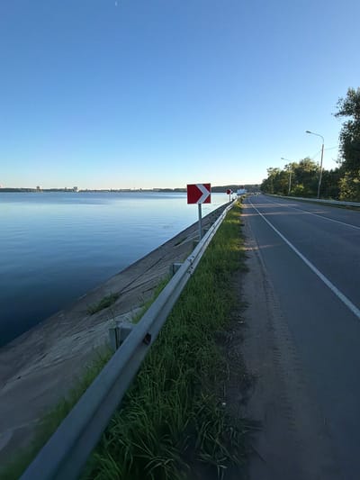 The image depicts a road running alongside a body of water, likely a lake or reservoir, with buildings visible in the distance across the water. The road has a white line marking the lanes and a guardrail separating it from the embankment. Green grass and trees line the roadside. Traffic signs, including a directional arrow sign indicating a curve, are visible. The sky is clear and blue with a faint crescent moon. The scene appears to be a scenic route, possibly near Solnechnogorsk, Russia, given the mention of a lake and typical infrastructure found in such locations.