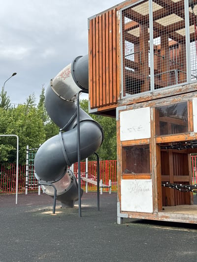 The image shows a playground structure with a wooden and metal tower that has a mesh grate on one side. Attached to the tower is a dark gray, curved slide. The slide appears to be made of plastic and has a metallic interior. There is some graffiti on the exterior of the slide. The playground is situated outdoors, with trees and a cloudy sky visible in the background. There are also red metal fences in the background, suggesting it is part of a larger park or recreational area. The ground is covered with a dark, textured surface, likely rubberized safety surfacing common in playgrounds. The overall scene suggests an overcast day. There are handwritten words visible on white panels on the wooden tower. One panel has the word "SPE" in large block letters. Another panel has cursive writing that is difficult to decipher but appears to be a name or message. A third panel also has cursive writing. The image is oriented vertically.