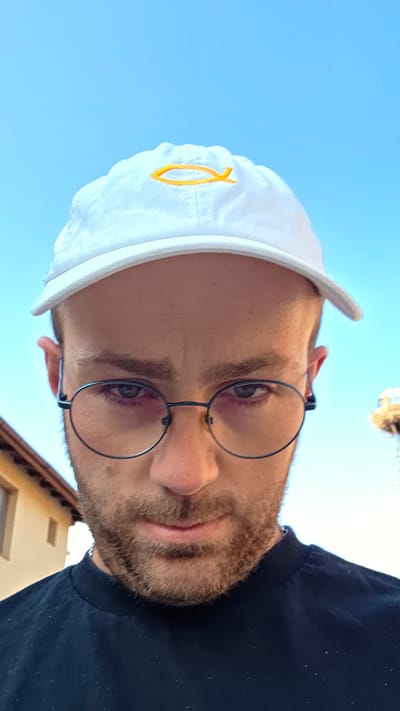 The photograph, taken in Bania, Bulgaria, shows a man from the chest up, looking directly at the camera. He is wearing a white baseball cap with a yellow fish symbol on the front and dark-rimmed, round glasses. His facial hair is short and reddish-brown. The background consists of a clear blue sky with a blurred building to the left and a tower-like structure on the right. The man is wearing a dark t-shirt.