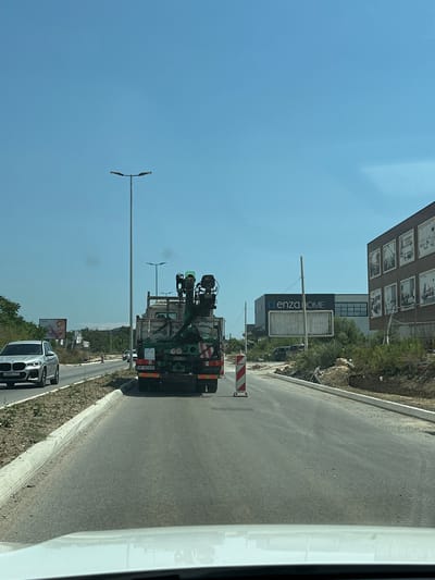 This image captures a daytime scene in Dub, Montenegro, likely during fair weather given the clear blue sky. The perspective is from inside a vehicle looking out. The primary focus is a street scene with ongoing construction or road work.

In the foreground, a large green truck with a crane attachment is prominently featured. It appears to be engaged in some form of heavy lifting or utility work, with its equipment extended. Traffic cones and barriers are visible, indicating a work zone. Further down the road, a silver BMW SUV is parked, its license plate visible.

Behind the construction, a modern building with a dark facade displays a prominent sign for "enza HOME." The building has multiple windows and appears to be a commercial establishment. In the distance, more trees and hills are visible, characteristic of a Mediterranean landscape. The road itself is asphalt and appears to be newly laid or under construction, with a concrete curb. Lampposts line the side of the road. The overall impression is one of a busy, developing area on a bright, sunny day.