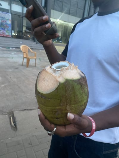 The image captures a close-up, dynamic view of a person holding a fresh coconut and a smartphone in an outdoor urban setting, likely in Ablekuma, Ghana. The subject, whose torso, arms, and hands are visible, is wearing a white t-shirt with dark accents and dark trousers.

In their right hand, the person holds a large, green coconut that has been expertly opened at the top, revealing the white inner flesh and the opening for consuming the coconut water. On their right wrist, a red silicone wristband is visible, bearing the clear white text "GOD IS HERE." A silver-colored ring with a dark stone is worn on the ring finger of their left hand. In their left hand, they hold a dark-colored smartphone.

The background shows a paved ground, possibly a sidewalk or street, and glimpses of modern buildings with reflective glass facades. A light-brown plastic chair is lying on its side in the mid-ground. The lighting suggests it is daytime. The overall scene depicts a casual moment of enjoyment, possibly on a break or while strolling.