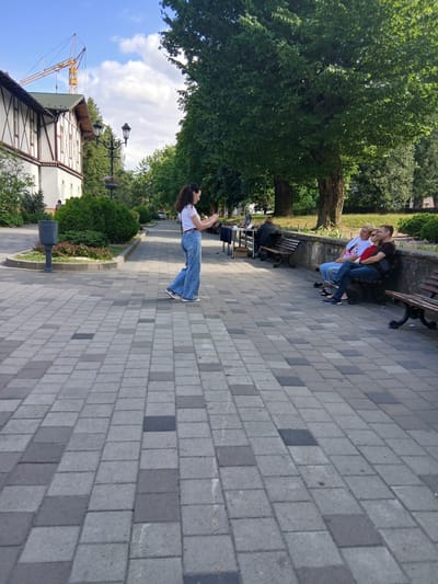 The image depicts a picturesque park scene in Truskavets, Ukraine, on a bright, sunny day with a few scattered clouds. The foreground is dominated by a meticulously paved walkway made of gray and darker gray square cobblestones, creating a visually appealing pattern.

To the left, a white building with distinctive dark brown trim and a green roof stands partially visible, suggesting a historic or well-maintained structure. Adjacent to the building is a dark gray trash receptacle and well-manicured green bushes. A tall lamppost with decorative elements is also present near the building. In the distance along the walkway, a yellow construction crane is visible against the blue sky, indicating ongoing development or renovation in the city.

The middle ground features a lush green park area with mature trees casting shadows on the ground. Several park benches are strategically placed along a low, weathered stone wall. On one of these benches, three individuals are seated: a woman with blonde hair wearing a white t-shirt and blue jeans, a woman with darker hair wearing a red top and blue jeans, and a man with dark hair wearing a black t-shirt and blue jeans. They appear to be engaged in conversation or simply enjoying the park. Another bench is positioned further down the walkway.

In the center of the walkway, a young woman with dark curly hair pulled into a ponytail is standing and looking at her phone. She is wearing a white top and stylish, wide-leg blue jeans. Her posture suggests she might be pausing or waiting.

The overall atmosphere of the image is serene and idyllic, capturing a moment of everyday life in a beautiful urban park setting.