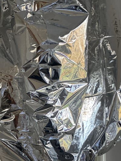 The scene, located in the city of Wealden, United Kingdom, consists of a vertical close-up of a severely crumpled, highly reflective silver material, consistent with aluminum foil or a similar metallic film. The material's surface is characterized by an intricate, chaotic network of sharp creases, deep folds, and irregular planar facets. This texture generates numerous bright highlights and dark, contrasting shadows, defining the material's three-dimensional relief. Reflections on the surface exhibit amorphous patches of bright white, yellow, and subtle green hues, indicating ambient light and potentially surrounding environmental elements, but no specific objects or figures are discernible within these reflections. There are no people, explicit objects beyond the crumpled material, actions, or interactions directly depicted within the frame.