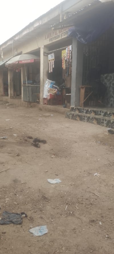 This image captures an unpaved outdoor scene in the city of Muhammadu Dikko Stadium, Nigeria. The primary focus is a single-story building with a faded, light-colored facade and a corrugated metal roof, viewed under an overcast sky. The central section features an open storefront labeled "A. MUH.IYAL MASJAWA PROVISION STORE" in dark, prominent lettering. Inside this store, an array of packaged goods hangs from strings, while large white and multi-colored sacks are stacked on red crates. A person is vaguely discernible within the store, partially obscured by these provisions. To the immediate left of the provision store, a red and white "MAGGI" branded umbrella shades a metal stand holding bottled beverages. Further left, another recessed building entrance is visible. To the right of the provision store, the building's lower facade is adorned with dark, patterned black and white tiles, leading to two steps. A small wooden stool stands near this entrance, where another individual is vaguely visible, seated or crouching behind a metal security grille. The foreground consists of dusty, uneven dirt and gravel, littered with crumpled black, white, and light blue plastic bags, along with other miscellaneous debris. No explicit actions or interactions are clearly depicted.