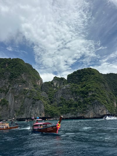 The image depicts a scenic view of limestone cliffs covered in lush greenery, characteristic of coastal Thailand. The sea is a deep blue, with visible waves and boat wakes, indicating movement and activity on the water. Several longtail boats, a common mode of transport in Thailand, are present. One boat in the foreground, painted with pink and blue accents, has Thai script and numbers on its hull. Another longtail boat to the left has "Adeeva" written on its side. In the background, more boats are visible, and a white speedboat is seen moving to the right. The sky is partly cloudy with a mix of white cumulus and wispy cirrus clouds. The overall impression is a vibrant and active marine environment set against a dramatic natural landscape.