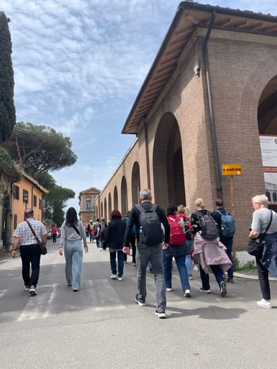 A group of tourists are walking down a paved road in Rome, Italy. They are located near the Basilica di Santa Sabina on the Aventine Hill, at the coordinates 41.884 latitude and 12.48 longitude. They are walking towards a brick building with arches, a sign reading "s.sabina", and a building that has a facade and a large window. The building appears to be a church, with a small garden on the left side of the road. The tourists are walking in a single file line, some of them are carrying backpacks, while others are holding olive branches. There are trees and bushes on either side of the road, and a blue sky with white clouds above. The environment is sunny. The tourists are all wearing casual clothing. Some of them are looking at their phones. The tourists are all walking in the same direction. There is a car parked on the left side of the road, and a person walking towards the camera on the right side of the road. The person on the right side of the road appears to be wearing a light-colored jacket and dark pants, and a handbag slung across their shoulder. The tourists are walking along the road, seemingly on a tour of the city.