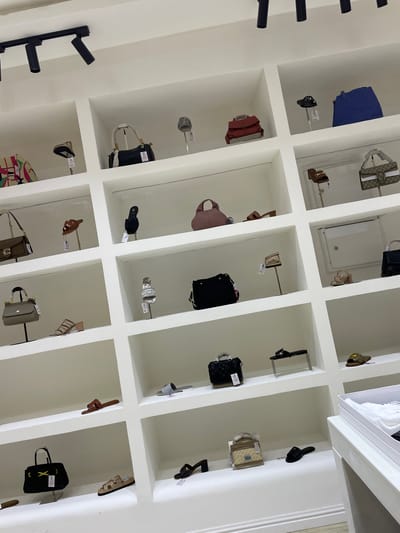 The image displays the interior of a retail store, likely a boutique, showcasing a collection of handbags and shoes arranged on white, recessed shelves. The store appears well-lit, with track lighting visible at the top of the frame.

On the top shelf, a red handbag is positioned centrally, flanked by a black hat on a stand to its left and a large blue tote bag to its right. The second shelf down features a black handbag with white handles and a decorative silver element. Below that, a pink handbag is displayed.

Further down, a collection of shoes is visible. On the third shelf from the top, a pair of brown sandals with a buckle is on display. On the fourth shelf, a taupe handbag with gold hardware is presented. A pair of beige, strappy heeled sandals is also visible on this shelf.

The fifth shelf from the top holds a black handbag with a white and red striped detail. Next to it, a pair of beige slides is showcased.

On the sixth shelf, a black quilted handbag is displayed on a stand. To its right, a pair of black slides is on a metal stand.

The bottom shelf shows a black handbag with a yellow clasp. Next to it, a pair of beige slides is placed. A single brown slide is also visible on this shelf, and another beige slide is to the right of it.

A white retail display box is partially visible on the far right of the image, containing white fabric items. The walls and shelves are painted white, creating a clean and minimalist aesthetic. The overall impression is one of a carefully curated selection of luxury fashion accessories. The location is identified as Unguwar Sarki, Nigeria.