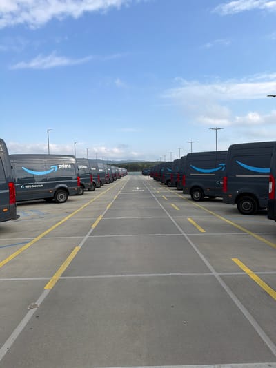 A vast fleet of dark grey Amazon Prime delivery vans is parked in neat rows within an expansive outdoor concrete lot. The image is taken from a slightly elevated perspective, looking down the length of multiple rows of vans stretching into the distance. Each van prominently features the Amazon "prime" text and the distinctive arrow logo in blue on its side. Many of the visible vans also display the text "100% Elektrisch" (100% Electric), indicating their power source, and some have individual numbering, such as "10," near the rear wheel.
The concrete pavement is clearly marked with a grid of yellow and white lines, defining individual parking spaces and pathways for vehicle movement. Overhead, the sky is bright blue with scattered white clouds, suggesting clear and pleasant daytime weather conditions. Tall light poles are visible throughout the parking area. The scene depicts a large-scale logistics or delivery hub, with the vans awaiting deployment or having just completed their routes. There are no people visible, and the vehicles are stationary. This scene is located in Friedrichsdorf, Germany.