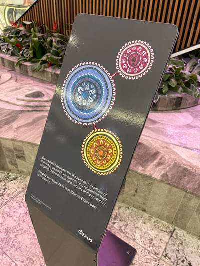 A black sign with a logo in white lettering that says "dexus" is positioned on the floor. The sign has three concentric circles in various colors and designs: one blue, one yellow, and one red.  The blue circle is on the left, the yellow is on the bottom, and the red is on the right. The circles are connected by dotted lines.  The sign is in a public indoor space with tiled floors. It is located at the Indooroopilly Shopping Centre, Indooroopilly, Australia (latitude: -27.5, longitude: 152.973). The sign has a text that reads "Dexus acknowledges the Traditional Custodians of the lands on which we operate and recognises their ongoing connection to land, waters and community. We pay our respects to First Nations Elders past and present." The sign is partially obscured by a small, purple object on the floor.  There are no people in the image.  The scene is lit by artificial lighting, and the lighting is uneven.