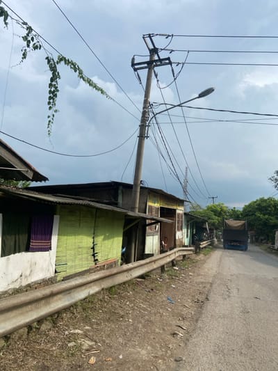 The image shows a street in Margaasih, Indonesia (latitude: -6.943, longitude: 107.556). The street is lined with small, simple houses with corrugated metal roofs. A large truck is driving down the street. The street is paved with asphalt, and the surrounding terrain is dirt. A utility pole with many power lines runs down the left side of the street. A streetlight is attached to the pole. A metal guardrail separates the street from the houses. There are several plants, vines and trees growing on the houses and the ground. The sky is cloudy.