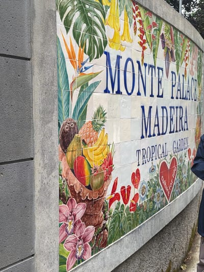 The image shows a tiled mural on a low platform in Funchal, Portugal. The mural depicts the words "MONTE PALACE MADEIRA TROPICAL GARDEN" in dark blue block letters, arranged vertically. The background of the mural is filled with colorful illustrations of tropical plants and fruits, including monstera leaves, yellow trumpet flowers, bird-of-paradise flowers, pineapples, bananas, mangoes, and orchids. A portion of a dark blue garment is visible in the upper right corner of the image. The platform surrounding the mural is made of gray concrete.