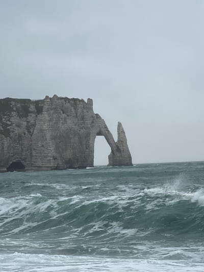 The image depicts the iconic cliffs and arch of Étretat, France, on a cloudy day. The sea in the foreground is rough, with waves crashing against the shore and the base of the cliffs. The cliffs themselves are tall and imposing, with layered rock formations and patches of green vegetation on top. A natural archway is visible in the central cliff, and a smaller rock formation stands to its right. The sky is overcast and a pale gray, contributing to the somewhat somber mood of the scene. No people or animals are visible.