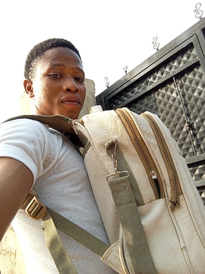 A medium-shot selfie shows a person with short, dark hair and brown skin wearing a white t-shirt and carrying a beige canvas backpack with two zippered compartments. The backpack has light-green straps and a metal clip. The person is positioned at an angle, looking directly at the camera. In the background, a dark-colored metal gate with a diamond pattern and decorative swirls is visible. The lighting suggests an outdoor setting, possibly during the day.