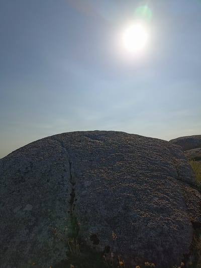 A wide-angle, low-angle shot captures a bright sun in the upper right corner of a clear blue sky. Below the sky, a large, textured granite boulder dominates the foreground, its surface appearing rough and speckled. A dark, thin crack runs down the center of the boulder. The base of the boulder is surrounded by dry grass and a few small, delicate wildflowers. To the right, another, less prominent boulder is visible, partially obscured by vegetation. The overall scene suggests an outdoor, natural environment, possibly a rocky outcrop or hillside. There are no people or animals present. The location is specified as Carnboy, Ireland.